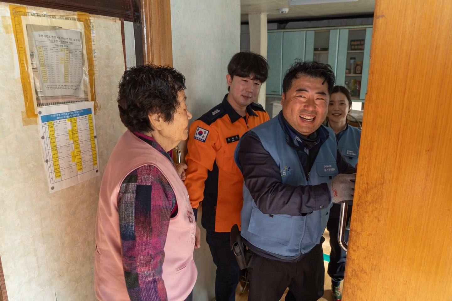 현관 문틀에 새로 단 손잡이 앞에서 웃는 집주인 할머니와 소방관, 봉사단 직원